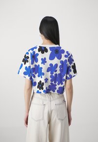 Mujer con cabello negro largo, vistiendo un top corto blanco con estampados de flores en azul y negro, y pantalones de tiro alto en un tono beige claro, de espaldas.