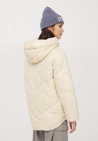 Puchowa kremowa kurtka z kapturem, z geometrycznym pikowaniem i błyszczącym wykończeniem. W zestawie z szarym dzianinowym beretem.