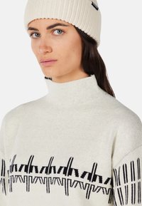 Light-colored knit sweater with a high collar, featuring a black geometric pattern along the bottom. The model wears a matching ribbed beanie.