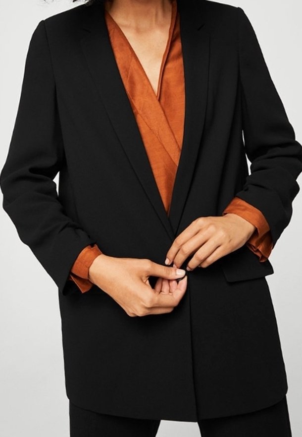 Personne portant un blazer noir sur une chemise couleur rouille, en train de boutonner le bouton avant du blazer avec les deux mains.