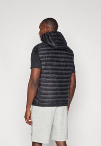 Črna napihljiva brezrokavnik s kapuco, z namensko quiltano zasnovo in gladko teksturo, oblečen čez črno majico s kratkimi rokavi in sivo kratke hlače.