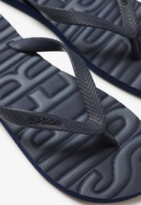 Tongs bleu marine avec semelle en caoutchouc texturée et brides à motif chevrons. Affiche une marque "SUPERDRY" sur la bride.