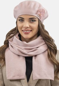 Világos rózsaszín kötött beret, textúrált felülettel és virágos díszítéssel; puha sál, amely a nyak köré van terítve.