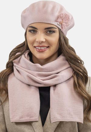Lyserød strikket beret med en tekstureret overflade og blomsterudsmykning; en matchende blød tørklæde draperet om halsen.