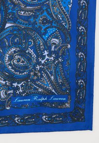 Μπλε ύφασμα με σχέδιο πεϊσλεϊ, διακοσμημένο με περίπλοκα μαύρα και λευκά λουλουδένια και σπειροειδή μοτίβα, που φέρει την υπογραφή "Lauren Ralph Lauren" σε καλλιγραφικό στυλ.