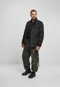Chaqueta negra de estilo militar con múltiples bolsillos, combinada con pantalones cargo de camuflaje y botas de combate negras, de pie contra un fondo neutro.
