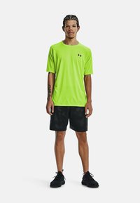 Felle limoengroene sportshirt met korte mouwen en een ronde halslijn, gecombineerd met zwarte shorts met een subtiel patroon en zwarte sneakers.