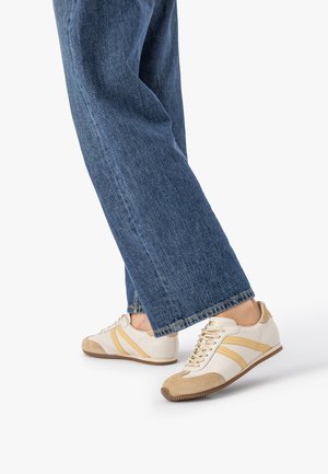 Osoba w niebieskich dżinsach i beżowo-białych trampkach z żółtymi paskami, jedna stopa lekko uniesiona na białym tle.