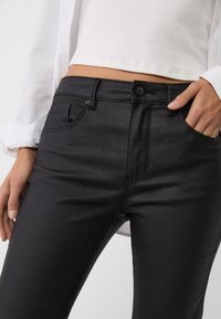 Zwarte denim broek heeft een gladde textuur, een klassiek vijf-pocket ontwerp en een knoopsluiting. Hand ligt op de heup, wat de slanke pasvorm benadrukt.
