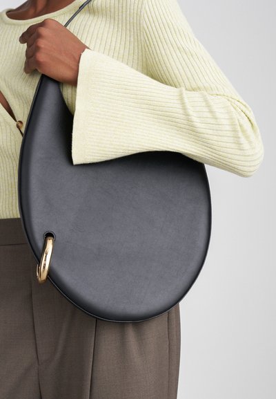 Apmēram melna ādas soma ar gludu tekstūru, ar zelta gredzena akcentu. Soma ir turēta pret vieglo riboto džemperi un brūnām biksēm.