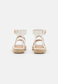 Bijela kožna sandala s otvorenom stražnjom stranom, dva bočna izreza i espadrille-stil razrađenim srednjim potplatom od jute. Ova sandala ima kopču na gležnju.