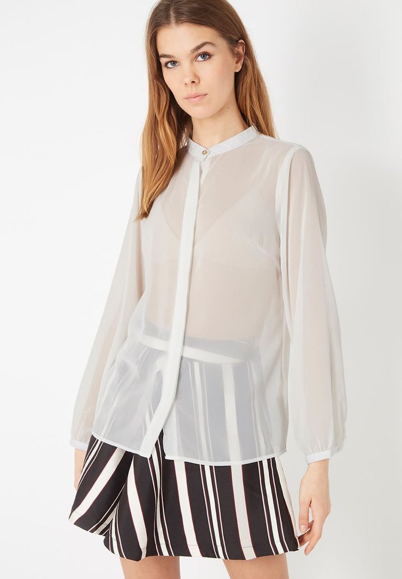 Blusa blanca transparente de manga larga, con cierre de botones y cuello redondo, combinada con una falda negra a rayas. Tela suave y ligera.