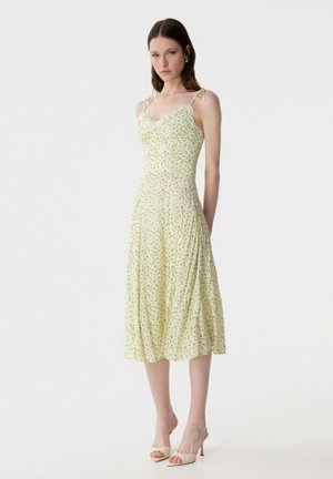 Giovane donna in piedi, indossa un vestito midi giallo chiaro con motivi floreali, spalline sottili e tacchi bianchi aperti, su sfondo bianco uniforme.