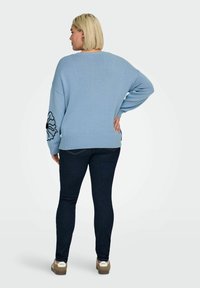 Maglione oversize azzurro chiaro con orlo a coste, decorato con un motivo floreale nero sulla manica, abbinato a jeans denim scuro e scarpe marroni.