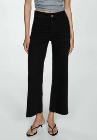 Pantalones vaqueros de mezclilla negros de pierna ancha con cintura alta, con un dobladillo sin terminar y bolsillos frontales. Combinados con sandalias negras de tiras.