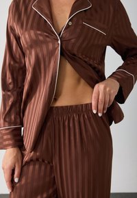 Ensemble de pyjama en satin brun avec des rayures verticales, une chemise à boutons avec une poche poitrine, et un pantalon à taille élastique au toucher doux.