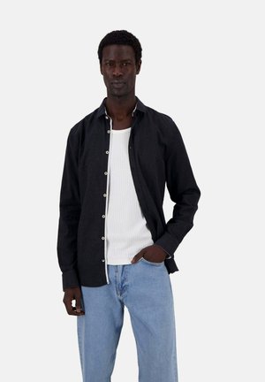 Schwarzes Langarmhemd mit Knöpfen und kontrastierendem weißen Nähten, getragen über einem weißen gerippten Tanktop und hellblauen, locker sitzenden Jeans.
