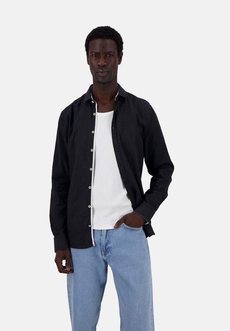 Schwarzes Langarmhemd mit Knöpfen und kontrastierendem weißen Nähten, getragen über einem weißen gerippten Tanktop und hellblauen, locker sitzenden Jeans.
