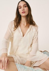 Joven con cabello largo y castaño, vistiendo una blusa traslúcida color crema con volantes y pantalones cortos a juego, sentada con las piernas abiertas sobre un fondo claro.