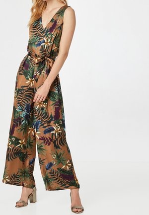 Femme portant une combinaison sans manches à col en V et jambes larges, base marron avec imprimé feuilles et fleurs tropicales vert, violet et crème, nouée à la taille.