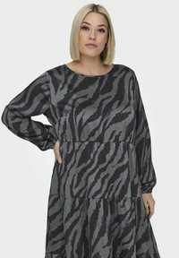 Harmaa ja musta polvipituinen mekko, jossa on tiikeriraidallinen kuvio, pitkät hihat ja kerroksellinen suunnittelu rentoa istuvuutta varten. Sujuva, teksturoitu kangas.