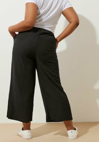 Svarta vida byxor med strukturerad yta, med midjeband och sidofickor, tillsammans med vita sneakers mot en neutral bakgrund.