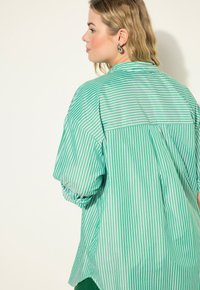 Camisa de gran tamaño a rayas verdes y blancas, con mangas enrolladas, dobladillo recto y panel en la parte posterior. Tela suave y ligera.