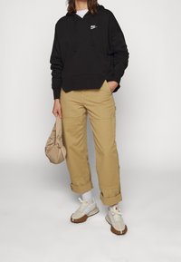 Zwarte katoenen hoodie met wit logo, beige cargobroek en witte sneakers met bruine zolen. Accessoire: lichtbruin geknoopte tas.