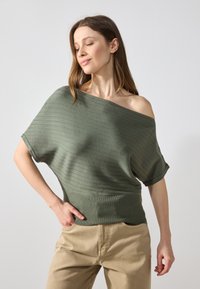 Pull en tricot côtelé ample de couleur verte douce, avec un décolleté épaule dénudée et des manches courtes, associé à un pantalon beige.