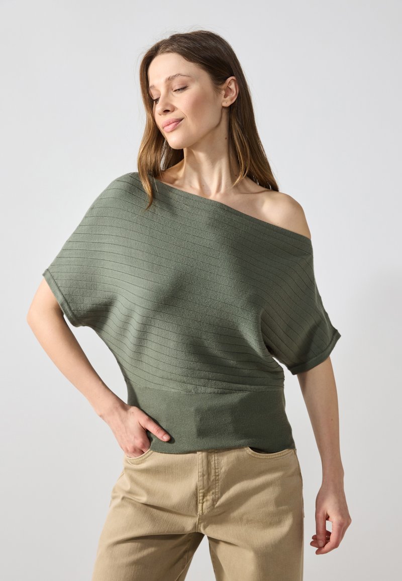 Pull en tricot côtelé ample de couleur verte douce, avec un décolleté épaule dénudée et des manches courtes, associé à un pantalon beige.