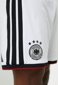 Λευκά ποδοσφαιρικά σορτς με μαύρες και άσπρες ρίγες, που διαθέτουν ένα στρογγυλό σήμα με έναν αετό και πέντε αστέρια από πάνω, με κόκκινες λεπτομέρειες.