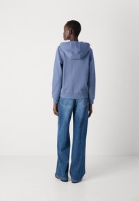 Un pull à capuche en tissu bleu doux, associé à un jean en denim à jambes larges. L'ensemble présente une coupe décontractée et un style minimaliste.