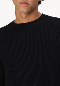 Juoda ripsuota megztinė su apvalia iškirpte ir tekstūruota medžiaga, matomi detalės kaklo ir pečių srityje. Priartinimas prie viršutinės dalies.