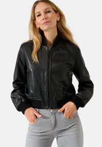 Giubbotto bomber in pelle nera con chiusura a zip, due tasche laterali, polsini a coste e vita aderente. Abbinato a jeans grigi.