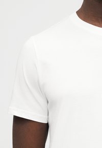 Gros plan d'une personne portant un t-shirt blanc uni à manches courtes sur un fond gris clair, montrant l'épaule et le haut du bras.