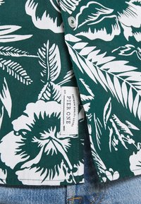 Vihreä kukkakuosiset paita, jossa on valkoisia lehtikuvioita, nappilista ja kudottu etiketti "PIER ONE" pohjahihnassa denimkankaan lähellä.