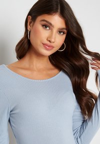Top a maniche lunghe blu chiaro a costine con scollatura ampia, tessuto testurizzato e design aderente. La modella mostra onde morbide nei capelli e orecchini a cerchio.