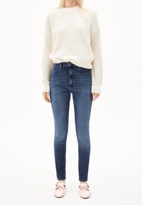 Tummansininen korkeavyötäröiset joustavat farkut, yhdistettynä väljään, tekstuuriin kermaiseen neulepuseroon ja vaaleanpunaisiin litteisiin kenkiin, joissa on solkikiinnitys.