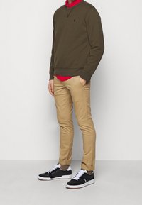 Felpa verde oliva, camicia rossa sotto, pantaloni kaki e sneakers nere con dettagli bianchi; vestibilità comoda, trama liscia e design casual.