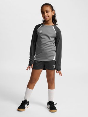 Ung pige iført gråt og sort sportstøj med hvide sokker og sorte sneakers på en ensfarvet hvid baggrund.