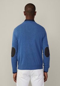 Sininen villaneule, jossa on tumma kyynärpäätuki, ribattuja hihansuita ja pyöreä pääntie, päällä tummansininen ruudullinen paita.