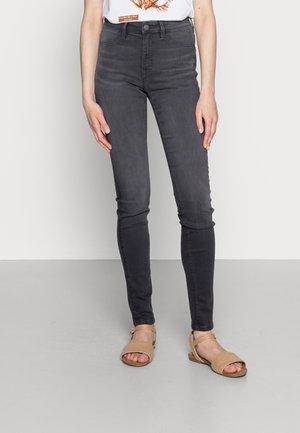 Person iført højtaljede mørkegrå skinny jeans og beige åbne sandaler, stående mod en hvid væg og grå gulv.