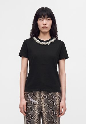 Femme aux longs cheveux noirs portant un haut noir à manches courtes avec un col décoratif en perles et un pantalon imprimé serpent, debout sur fond blanc.