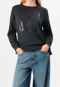 Henkilö, joka pitää yllään mustaa GAP-hupparia kiiltävillä mustilla kirjaimilla ja siniset farkut, kädet edessä taskuissa.