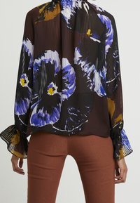 Schwarze, plissierte Bluse mit großen lila und gelben Blumenmustern, langen Ärmeln mit Rüschensäumen und einem lockeren Schnitt.