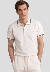 Bež majica polo s ovratnikom i manžetama u tamnoplavoj prugastoj boji, s malim logom na prsima. Nosena s bež hlačama.
