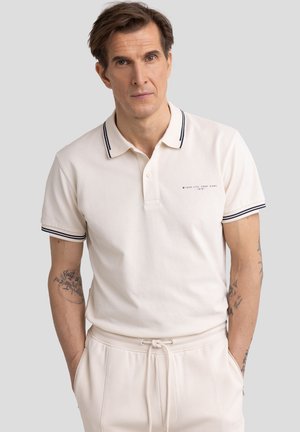 Beige Poloshirt mit navy-blau gestreiftem Kragen und Manschetten, versehen mit einem kleinen Logo auf der Brust. Kombiniert mit beigen Hosen.