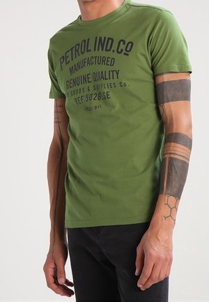 Person trägt grünes T-Shirt von Petrol Ind. Co und schwarze Hose, zeigt tätowierte Arme mit geometrischen und einfarbig schwarzen Designs.