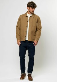 Chaqueta de algodón color cáñamo con cierre de botón, dos bolsillos frontales y un sutil acento de logo. Combinada con vaqueros oscuros y botas de cordones marrones.