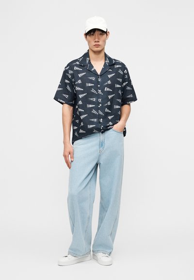 Νεαρός άνδρας που φοράει μπλε σκούρο πουκάμισο με λευκά γραφικά, ανοιχτό μπλε παντελόνι ευρύ μοτίβο, λευκά αθλητικά παπούτσια και λευκό καπέλο μπέιζμπολ, στέκεται με το ένα χέρι στην τσέπη.